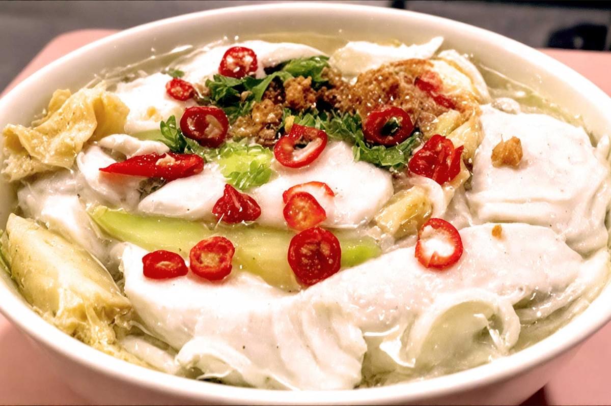 A white bowl is filled with fish slices, green vegetables, and garnished with sliced red chilies and herbs. The dish exudes a fresh and spicy look.