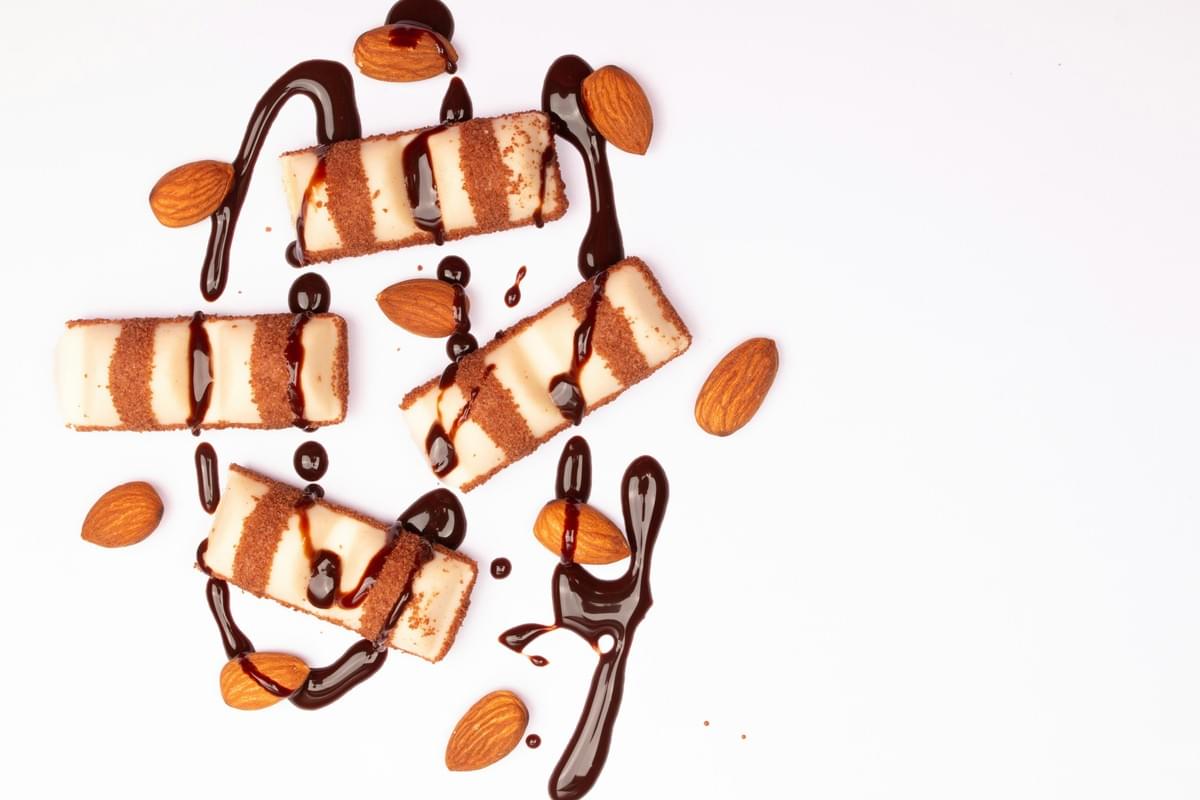 An overhead shot shows four white chocolate-coated wafer bars arranged on a clean white surface, drizzled with dark chocolate syrup. Whole almonds are scattered around the bars, adding a contrasting texture to the sweet dessert arrangement.