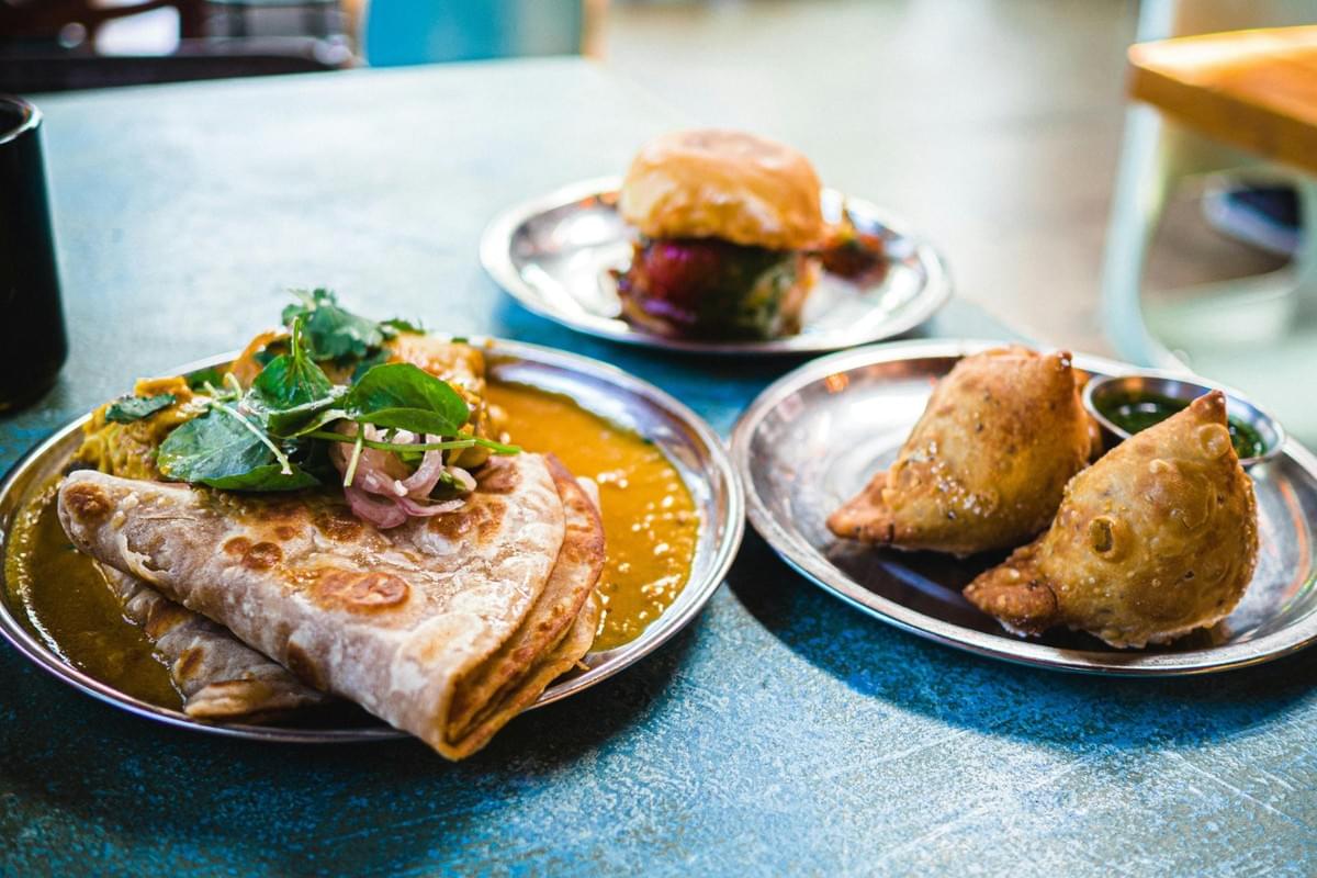 A table spread features three metal plates filled with Indian-inspired dishes. On the left, folded flatbread sits alongside a curry garnished with fresh greens, while two golden samosas with dipping sauce and a small slider burger occupy the plates on the right.