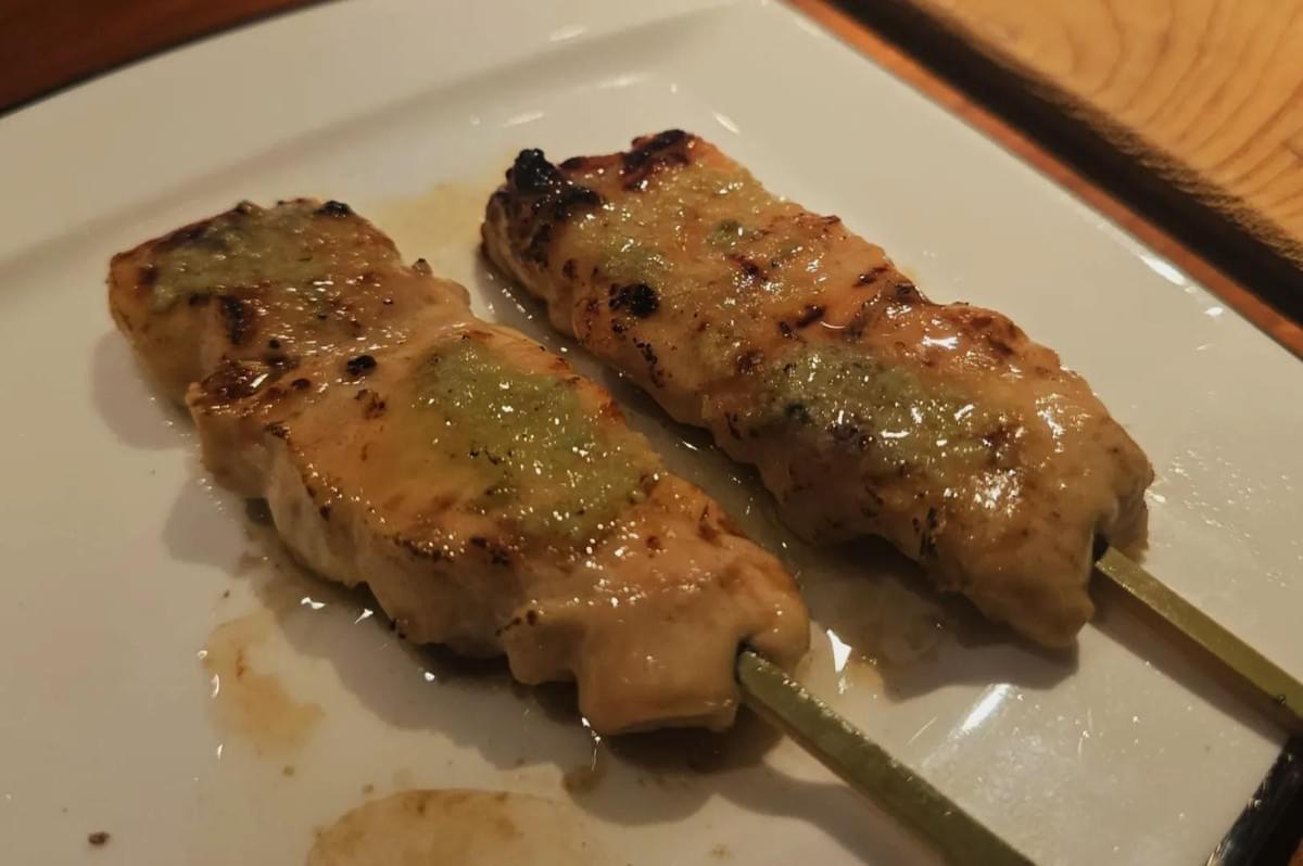 The image features two glossy, grilled chicken yakitori skewers resting side-by-side on a white square plate. Each skewer is lightly charred on the edges and topped with a stripe of green wasabi paste.