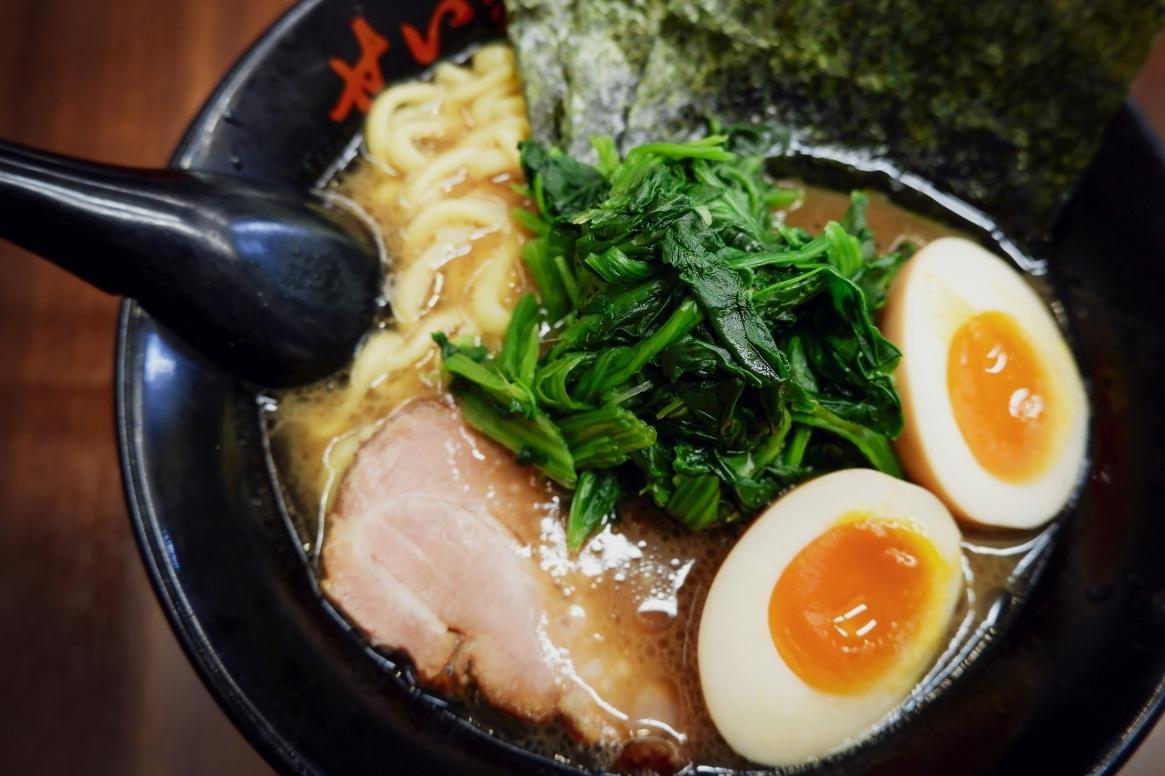 A close-up view shows a steaming bowl of ramen served in a dark ceramic bowl. The dish features thick noodles in a savory broth, topped with a generous mound of vibrant green spinach, a slice of chashu pork, two soft-boiled egg halves with jammy yolks, and a sheet of nori seaweed.