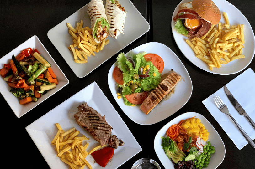 A bird's-eye view of a black table covered with a diverse spread of finished restaurant meals on square and round white plates, including grilled steak with fries, a salmon fillet with a side salad, a vibrant poke bowl, and chicken wraps.