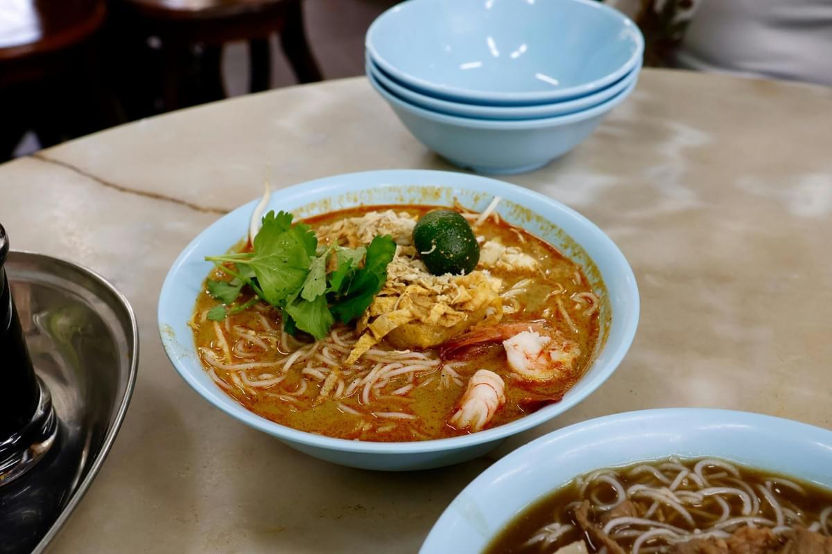 A steaming bowl of laksa is presented in a light blue bowl on a marble tabletop, garnished with fresh herbs, a calamansi lime, and succulent prawns.