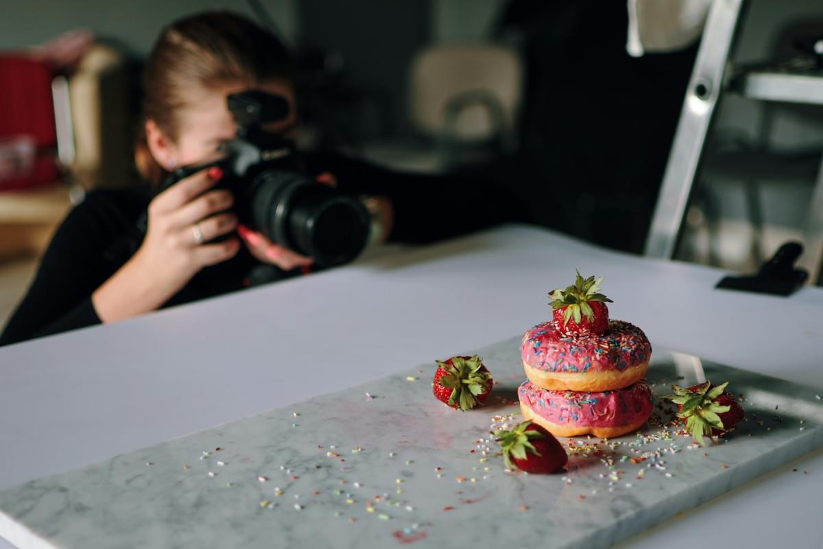 A stacked pair of pink-glazed donuts with sprinkles and fresh strawberries rests on a marble slab during a food photoshoot. In the blurred background, a photographer aims a professional camera to capture the dessert arrangement.