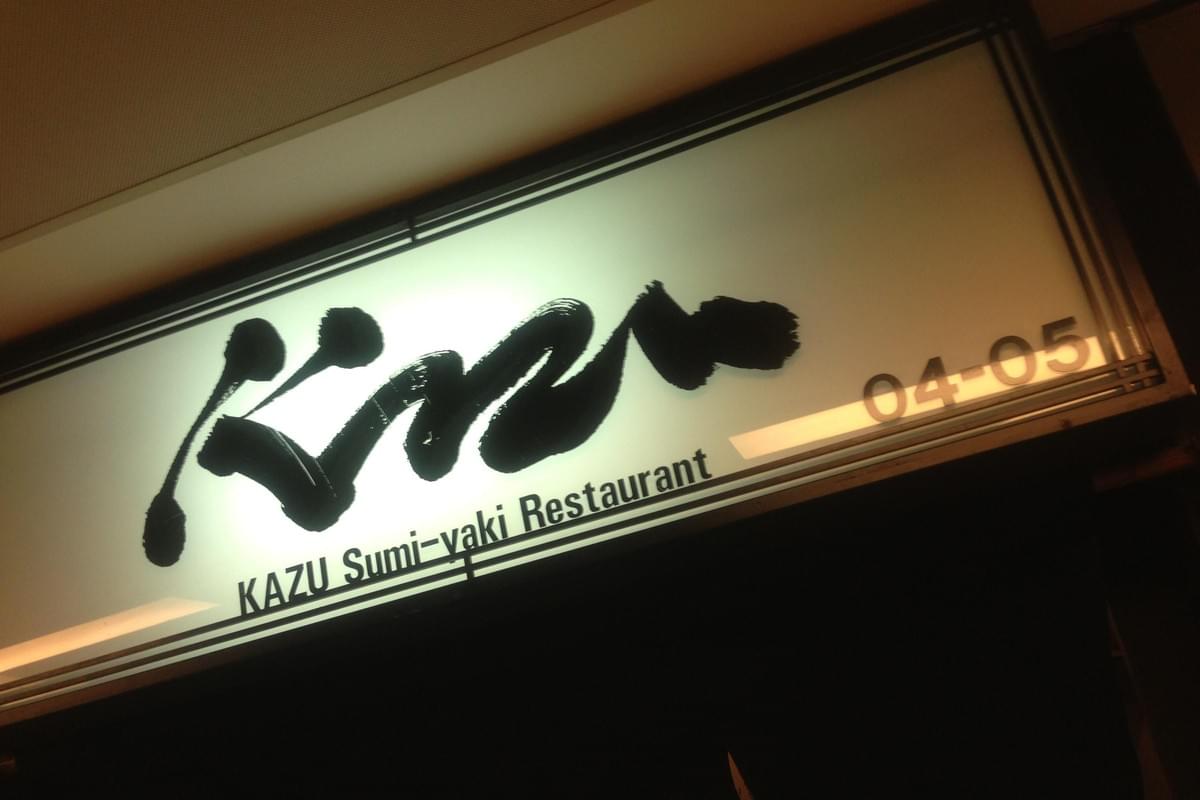 A low-angle shot shows a glowing restaurant sign featuring a stylized, calligraphic black logo that resembles the word "Kazu" written with artistic brushstrokes. Below the logo, the text reads "KAZU Sumi-yaki Restaurant" next to the numbers "04-05" on the right side.