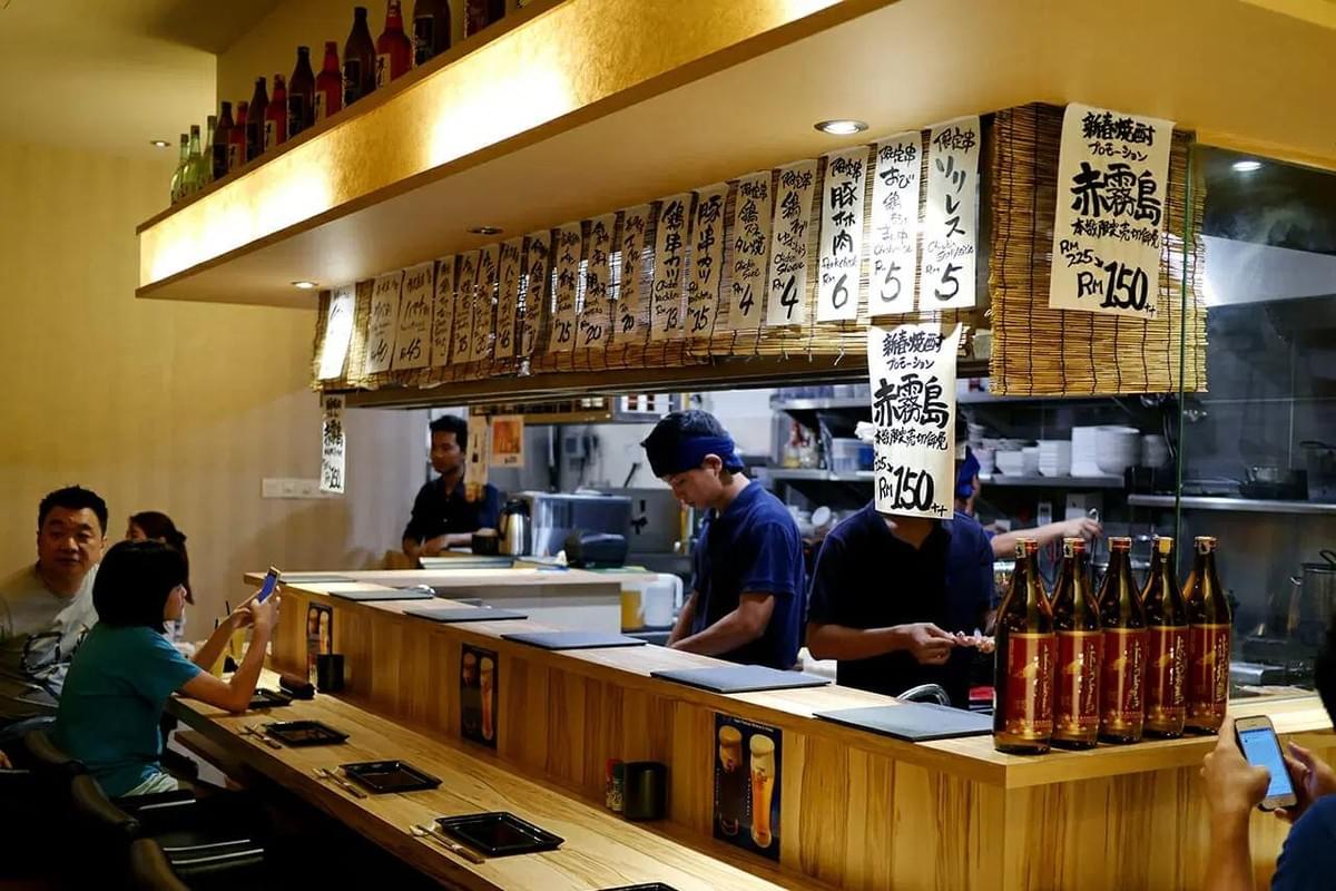 The image shows the interior of a modern Japanese restaurant featuring a light-colored wooden sushi counter lined with black bar stools. Several staff members are visible behind the counter and in the hallway, working under warm, ambient lighting.