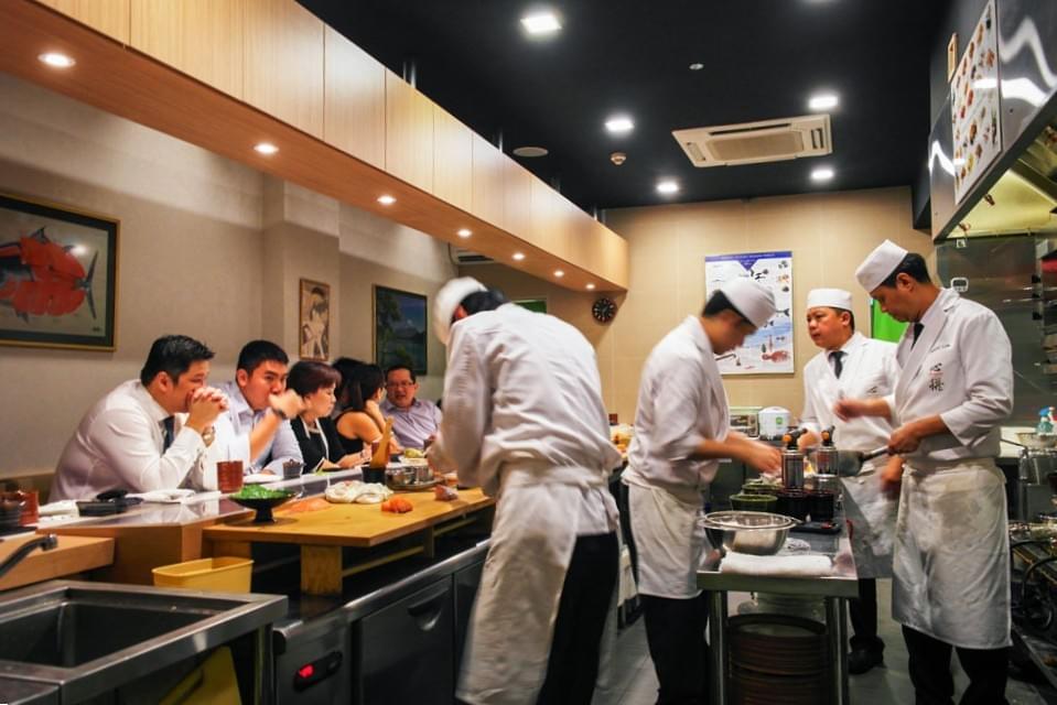 The image shows a busy open kitchen where chefs in white uniforms prepare fresh sushi behind a sleek wooden counter. In the background, diners sit closely together in a modern, brightly lit restaurant atmosphere.