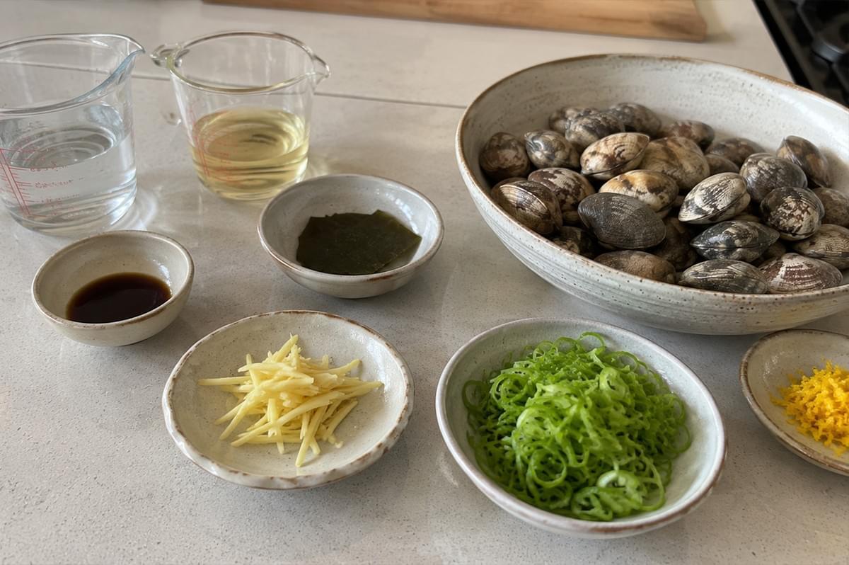 An organized mis en place layout is displayed across a white kitchen counter, showcasing all the raw ingredients needed for the recipe. A large, shallow ceramic bowl on the right is piled high with closed, uncooked clams in striped shells. Surrounding it are smaller matching ceramic dishes containing julienned fresh ginger, finely minced bright green scallions, dark brown soy sauce, a square piece of dried kombu kelp, and fine yellow citrus zest. Two clear glass measuring cups stand in the upper left, one filled with water and the other with a pale yellow sake or mirin liquid.