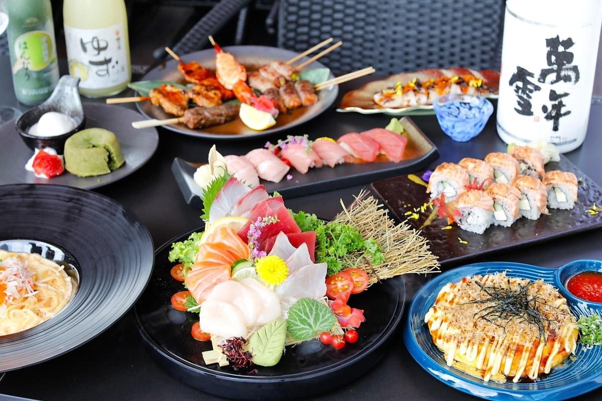 A vibrant spread of Japanese cuisine features a central plate of fresh sashimi alongside platters of sushi rolls, nigiri, and skewered yakitori. The table is also filled with a savory okonomiyaki pancake, a pasta dish, matcha dessert, and bottles of sake.