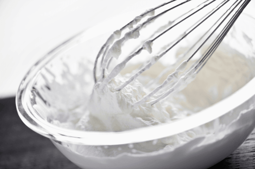 A close-up of a metal whisk lifting stiff peaks of freshly whipped meringue from a glass mixing bowl—highlighting its glossy texture and airy structure.