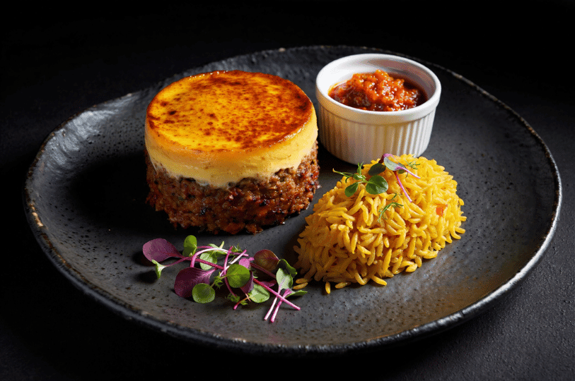 Gourmet presentation of a circular molded bobotie with a perfectly browned custard top, paired with a mound of yellow rice and microgreens on a dark plate.
