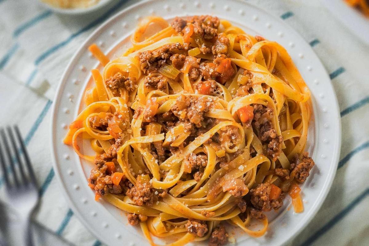 Italian tagliatelle pasta al ragù with tomato paste, ground meat, carrots, celery, onions, and grated cheese on white plate.
