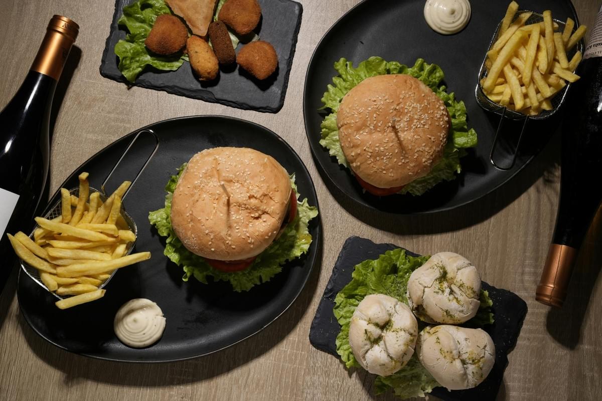 A top-down view shows a meal spread with two sesame seed burgers, crispy French fries, mayonnaise, and fried appetizers served on dark plates. Two wine bottles frame the wooden table, completing the dining scene.
