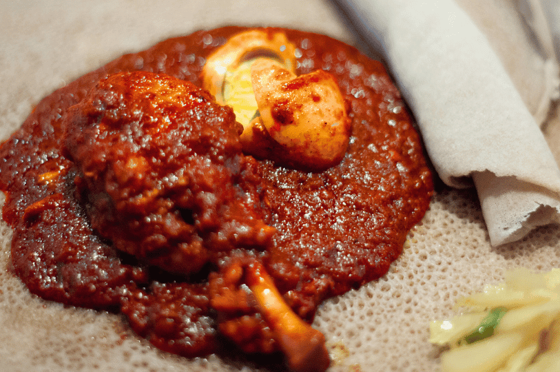 A close-up of Doro Wat served on a bed of porous injera bread, featuring shredded chicken and a sauce-coated egg next to rolled injera.