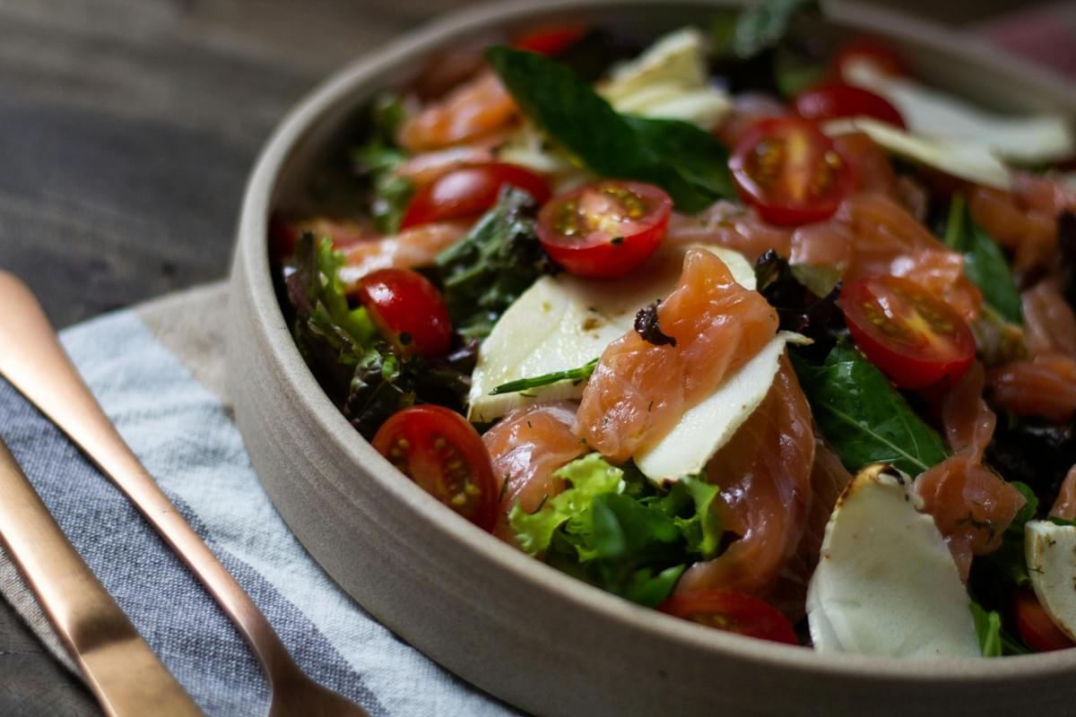 This fresh salad bowl features folded slices of smoked salmon, halved cherry tomatoes, and wedges of white cheese or fruit arranged over a bed of mixed greens. The dish is presented in a shallow beige bowl resting on a wooden table, accompanied by copper-colored cutlery and a striped napkin.