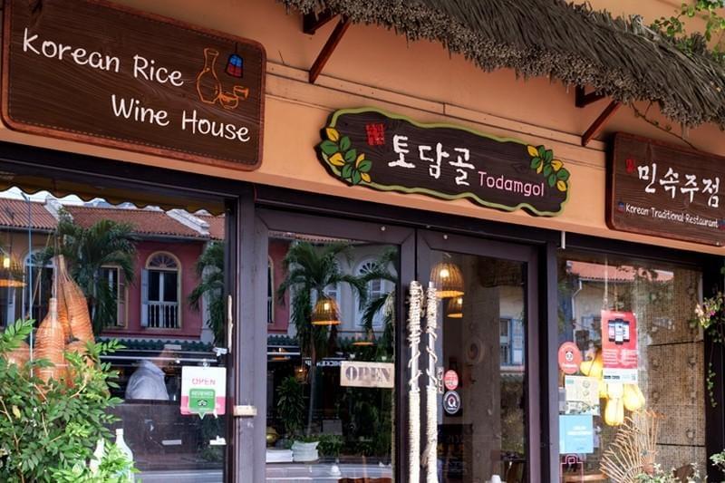 The image features the storefront of a Korean restaurant called "Todamgol," also labeled as a "Korean Rice Wine House." The entrance has dark-framed glass doors, wooden signage written in both English and Korean script, and a partial thatched roof accent above the signs.