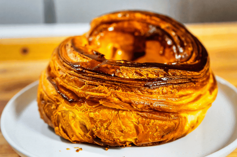 A close-up of a Kouign-amann, a caramelized multi-layered buttery pastry from Brittany, sitting on a white plate. The image captures the deep golden-brown, sugary crust and the flaky, laminated layers of the dough.