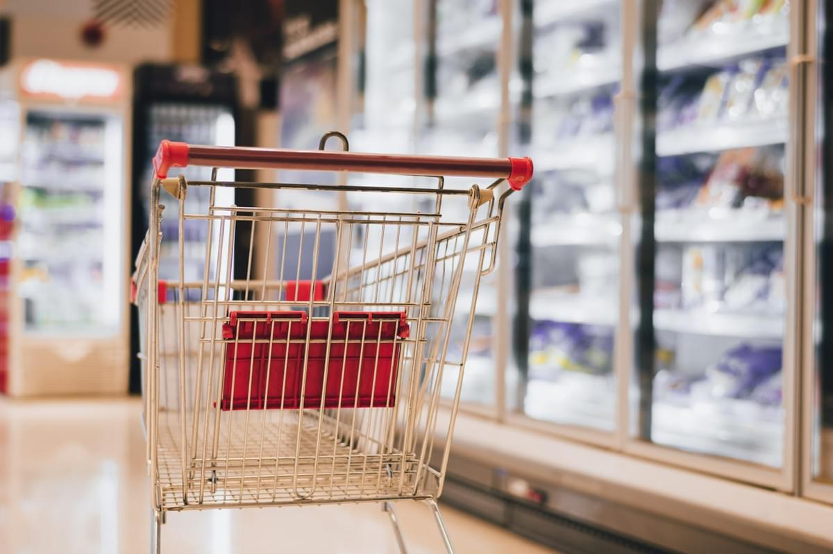 Empty shopping cart inside a grocery store aisle with bright refrigerated shelves in the background.