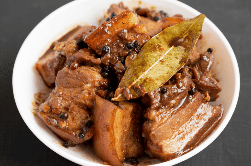 A white bowl filled with Filipino Pork Adobo featuring tender chunks of pork belly in a dark soy-vinegar sauce, topped with whole black peppercorns and a single dried bay leaf.