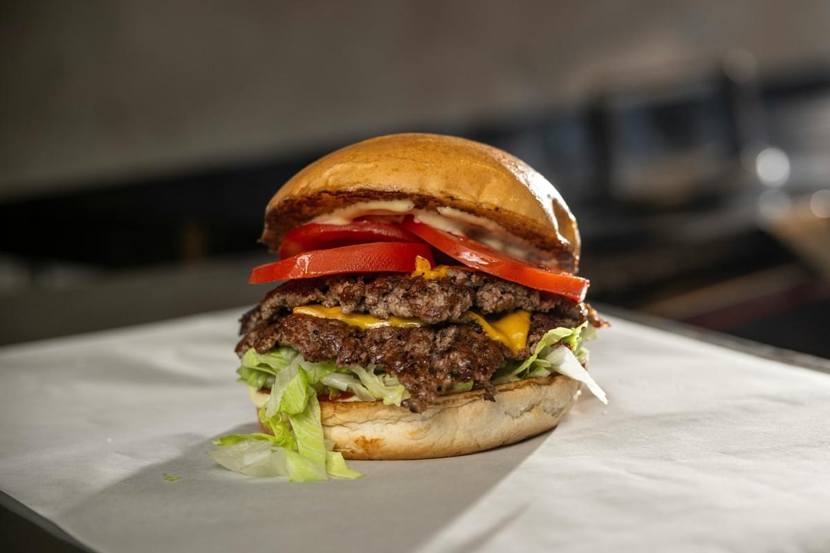 A double cheeseburger with two smash patties, melted yellow cheese, thick tomato slices, shredded lettuce, and sauce is served on white paper. The burger is held together by a toasted brioche bun against a blurred kitchen background.