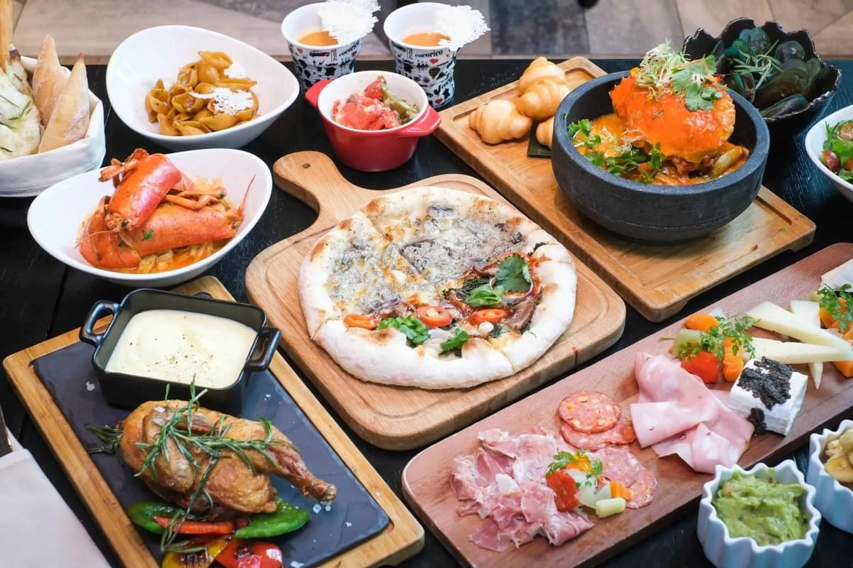 An overhead view shows a table densely covered with a lavish Italian dining spread. The feast includes roasted chicken, sliced charcuterie, fresh pizza, pasta dishes, lobster, and various appetizers.