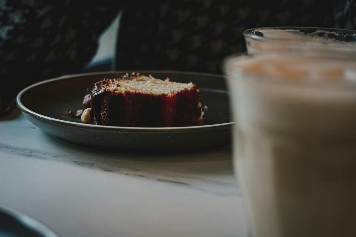 A thick slice of pound cake served on a dark gray plate sits in focus on a light-colored table. In the foreground, a tall glass filled with a frothy iced latte or coffee drink is partially blurred.