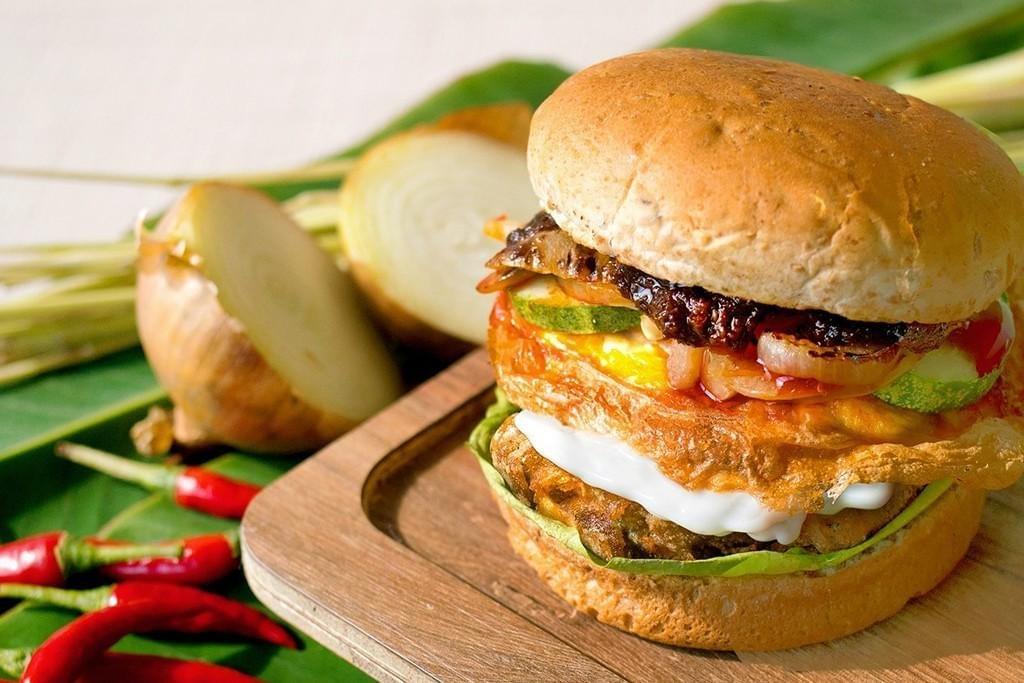 A stacked plant-based burger sits on a wooden board, layered with a vegan patty, fried egg alternative, cucumber slices, and a dark chili jam. The background features fresh culinary ingredients including vibrant red chilies, sliced onions, and aromatic lemongrass stalks on a green leaf.