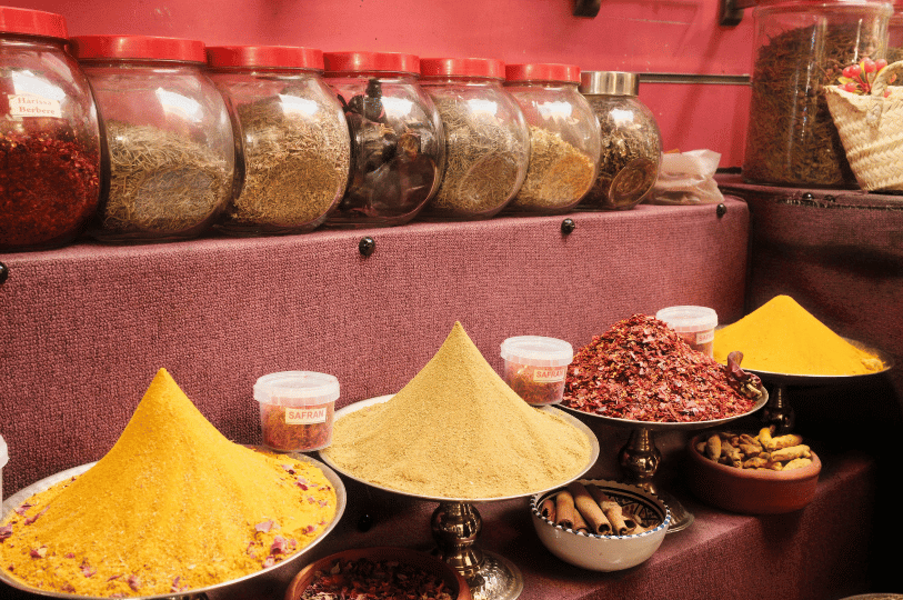 An indoor spice shop featuring large glass jars of dried herbs on a shelf above elegant, conical mounds of ground spices like turmeric and cumin displayed on silver stands, with bowls of cinnamon sticks and saffron nearby.