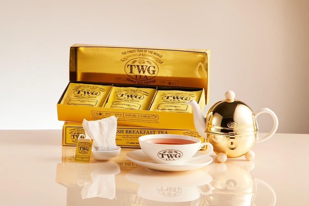 An open, metallic gold TWG Tea gift box displays three individually packaged tea sachets alongside a matching gold teapot and teacup set. A single cotton teabag rests in a small dish next to a freshly brewed cup of tea on a reflective surface.