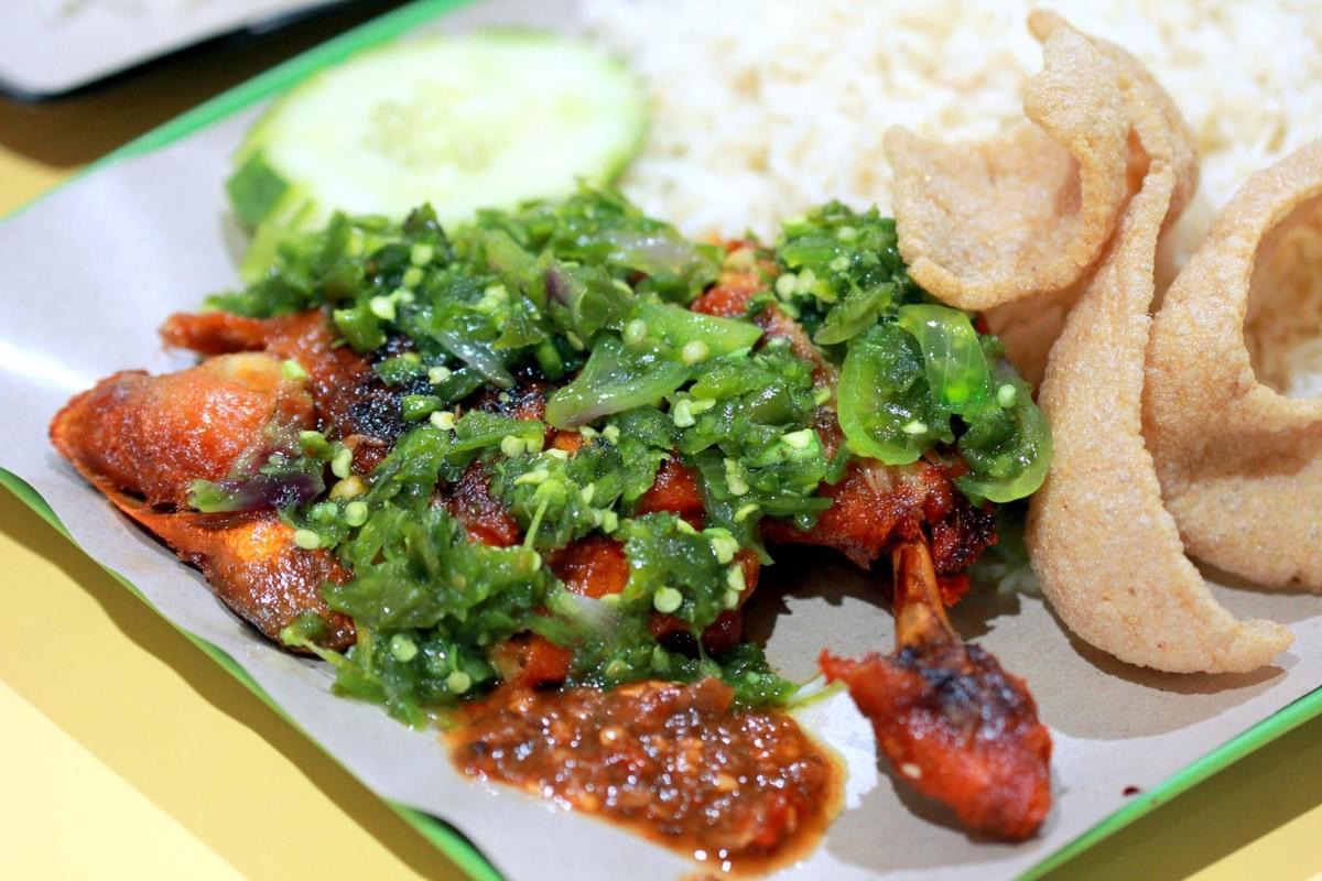 A piece of crispy fried chicken leg is topped with a generous layer of green chili sambal and served alongside spicy red chili paste. The meal is accompanied by white rice, crispy crackers, and a slice of fresh cucumber on a lined platter.