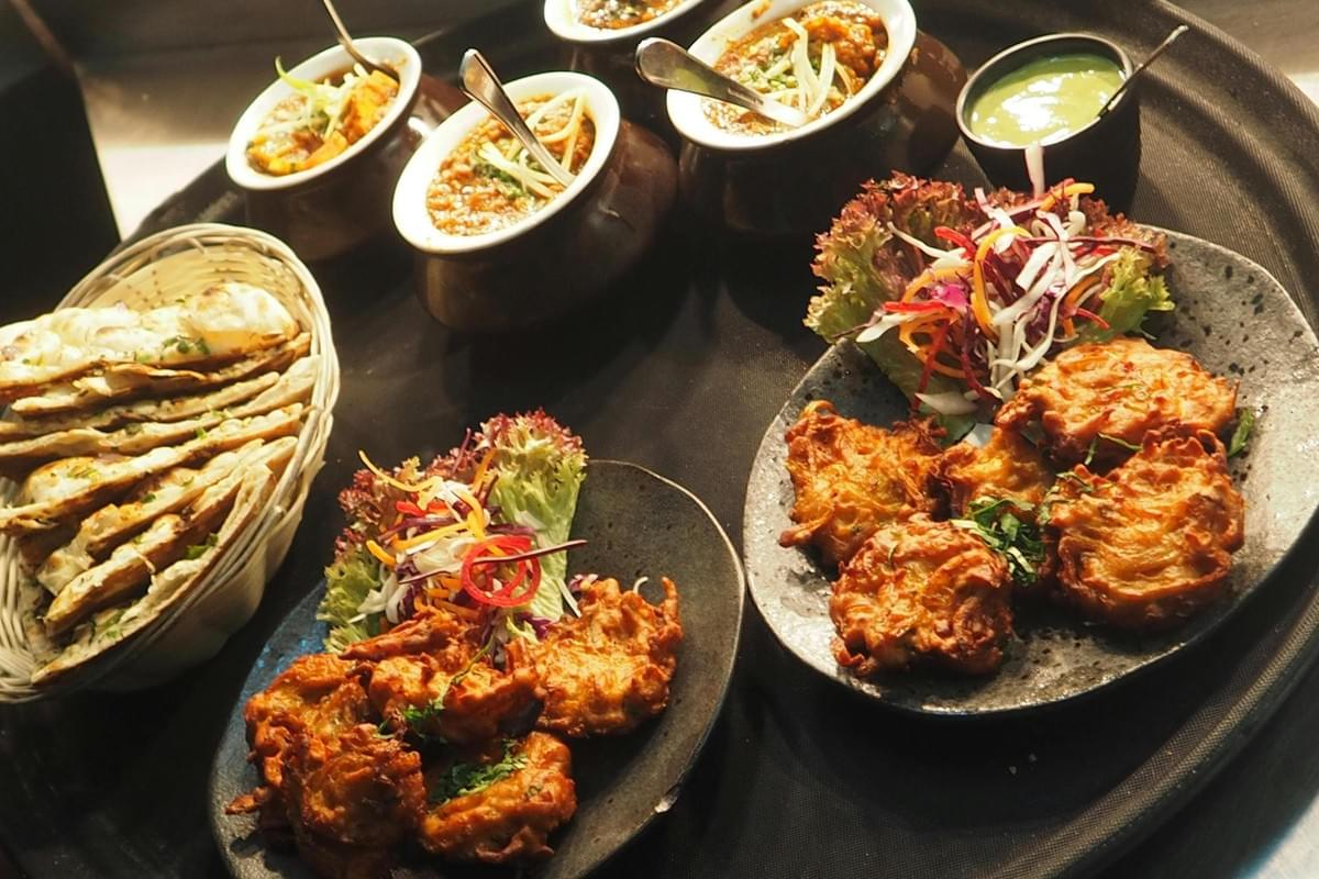 An assortment of Indian dishes includes small brown pots of curry with spoons, a basket of naan, and plates of golden-brown pakoras served with fresh salad. A small bowl of vibrant green chutney sits in the background on the black serving platter.