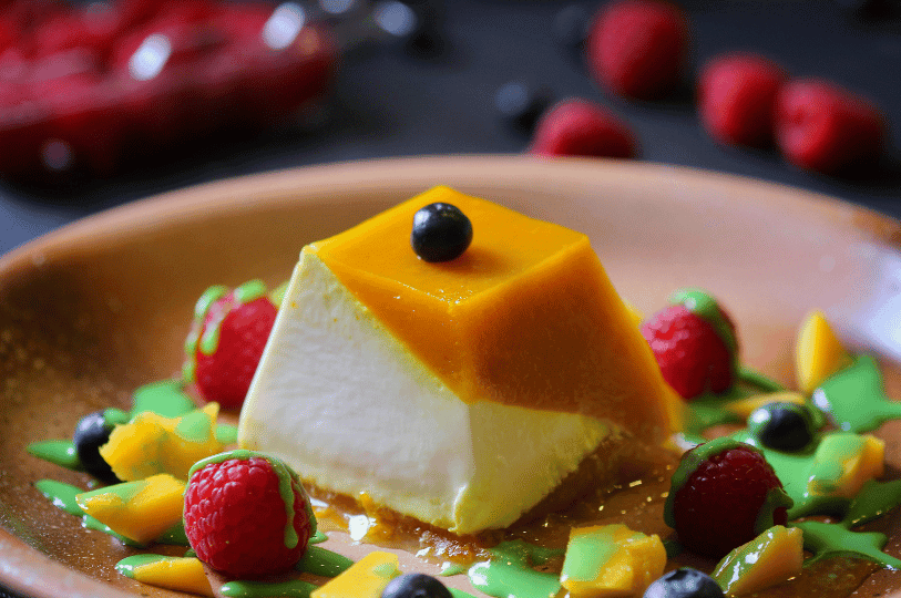 A sophisticated square-shaped Panna Cotta topped with bright mango glaze, served with fresh raspberries, blueberries, and a decorative drizzle of lime-green sauce.