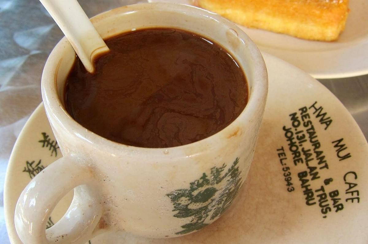 A cup of dark coffee with a white spoon rests on a saucer at Hwa Mui Cafe, partially showing their logo. A slice of toast is in the background.