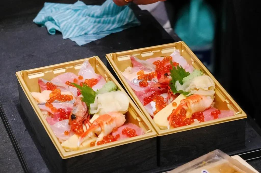 Two gold-bordered bentos sit side by side, generously filled with fresh sashimi, raw shrimp, crab claws, and salmon roe over rice. Green shiso leaves, wasabi, and pickled ginger are neatly placed alongside the colorful seafood toppings.