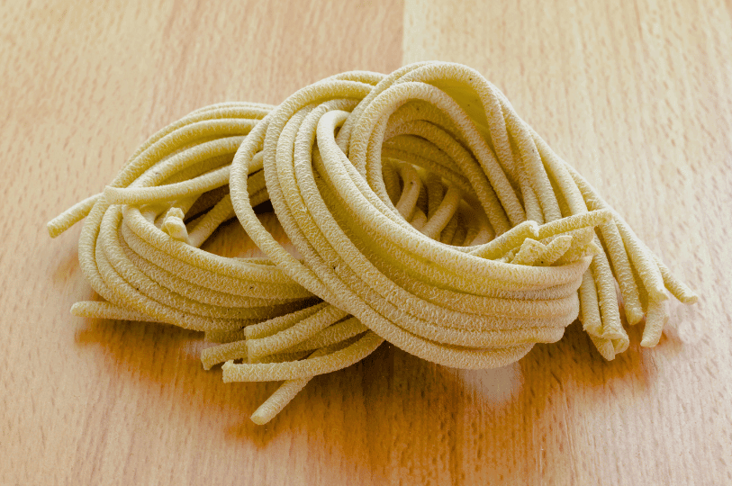 Two nests of thick, hand-rolled pici pasta, a rustic, spaghetti-like shape originating from Tuscany, resting on a light wooden surface.