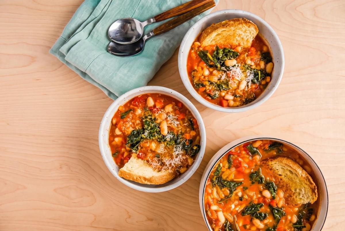 Three ceramic bowls of warm Tuscan white bean and kale soup served with toasted bread.