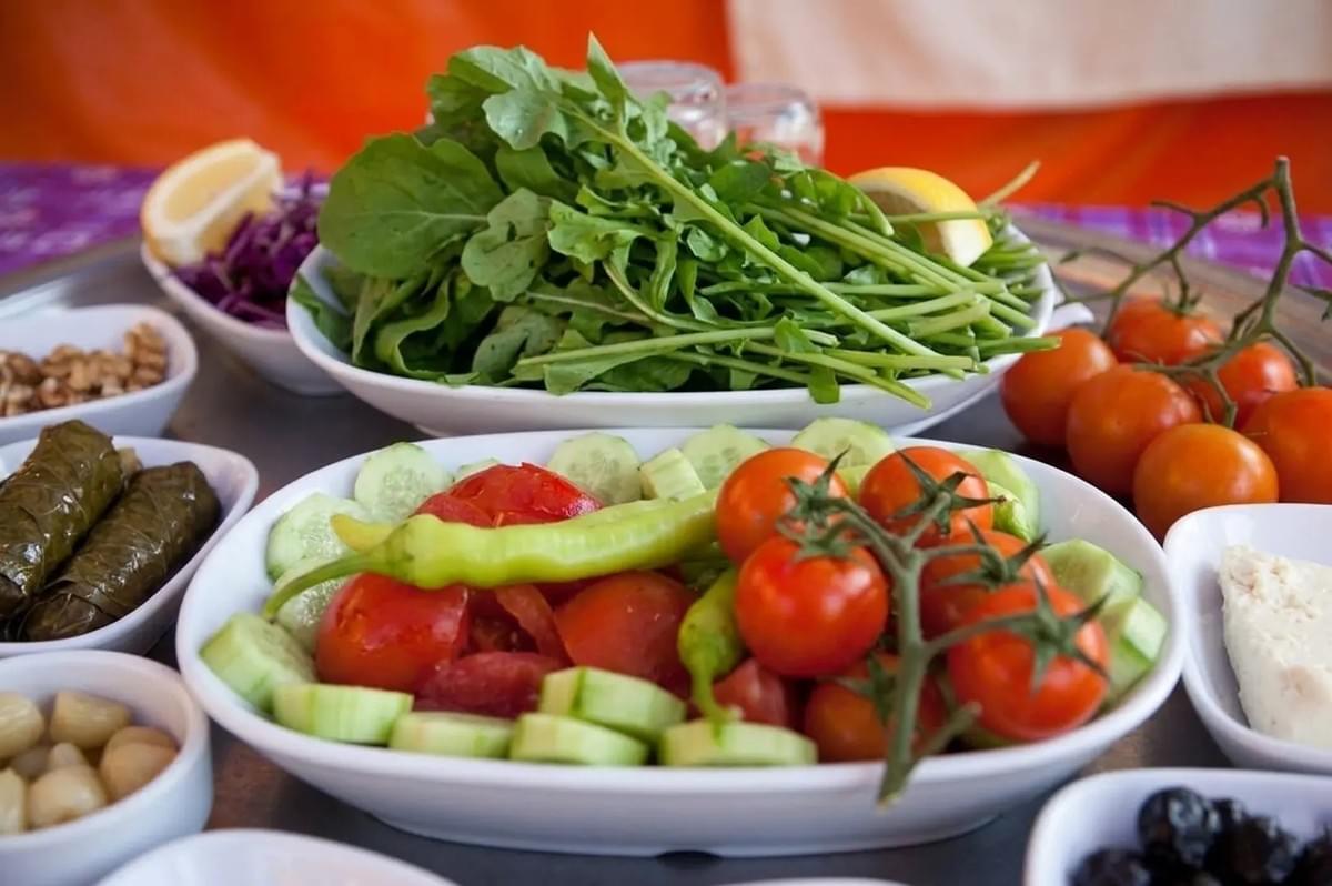 Colorful Turkish breakfast spread featuring fresh tomatoes, cucumbers, peppers, arugula, stuffed grape leaves, and olives in white bowls