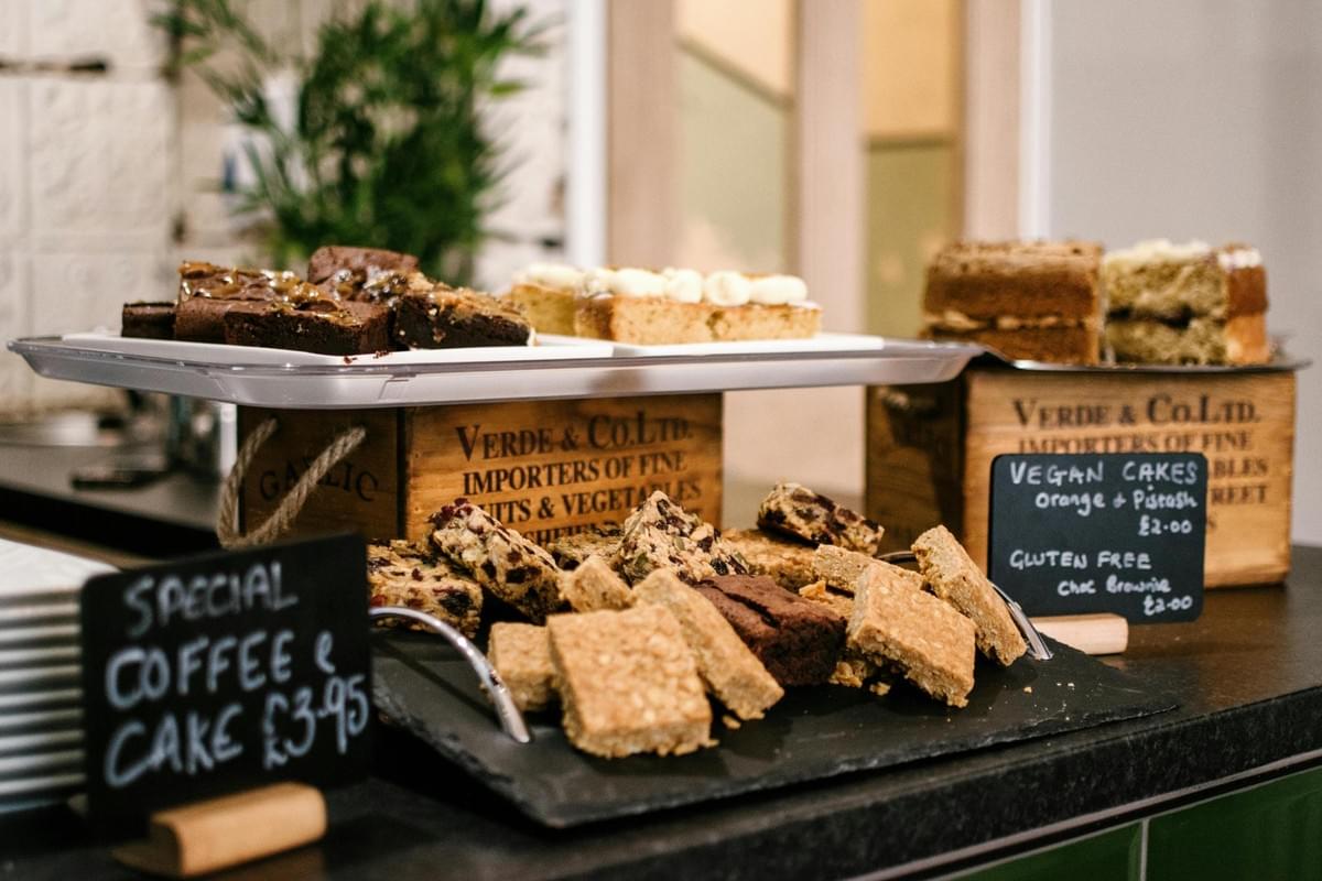 A tempting array of baked goods, including rich chocolate brownies, frosted cake slices, and golden oat bars, is displayed on tiered platters supported by rustic wooden crates. Handwritten chalkboards sit nearby, advertising a coffee special and identifying specific dietary options like vegan and gluten-free treats.