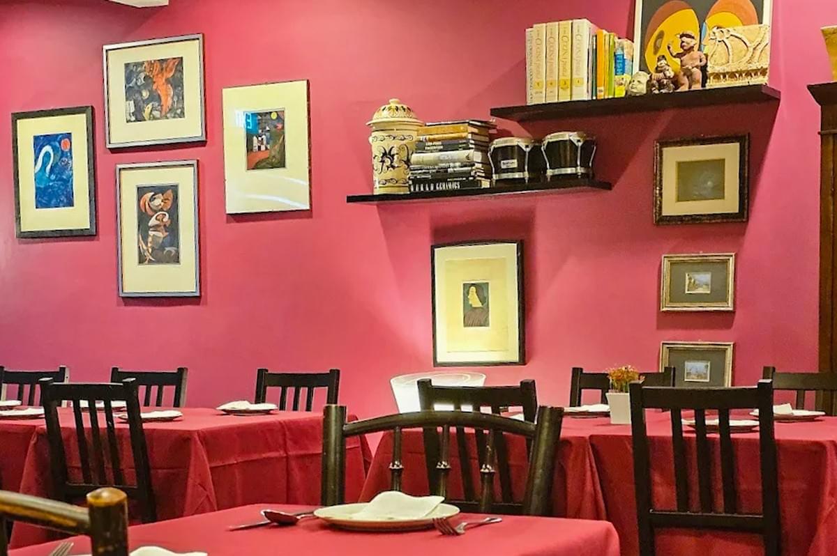 Interior view of a restaurant with deep red walls, multiple framed artworks, and floating shelves displaying books and decorative items above dining tables set with red tablecloths.
