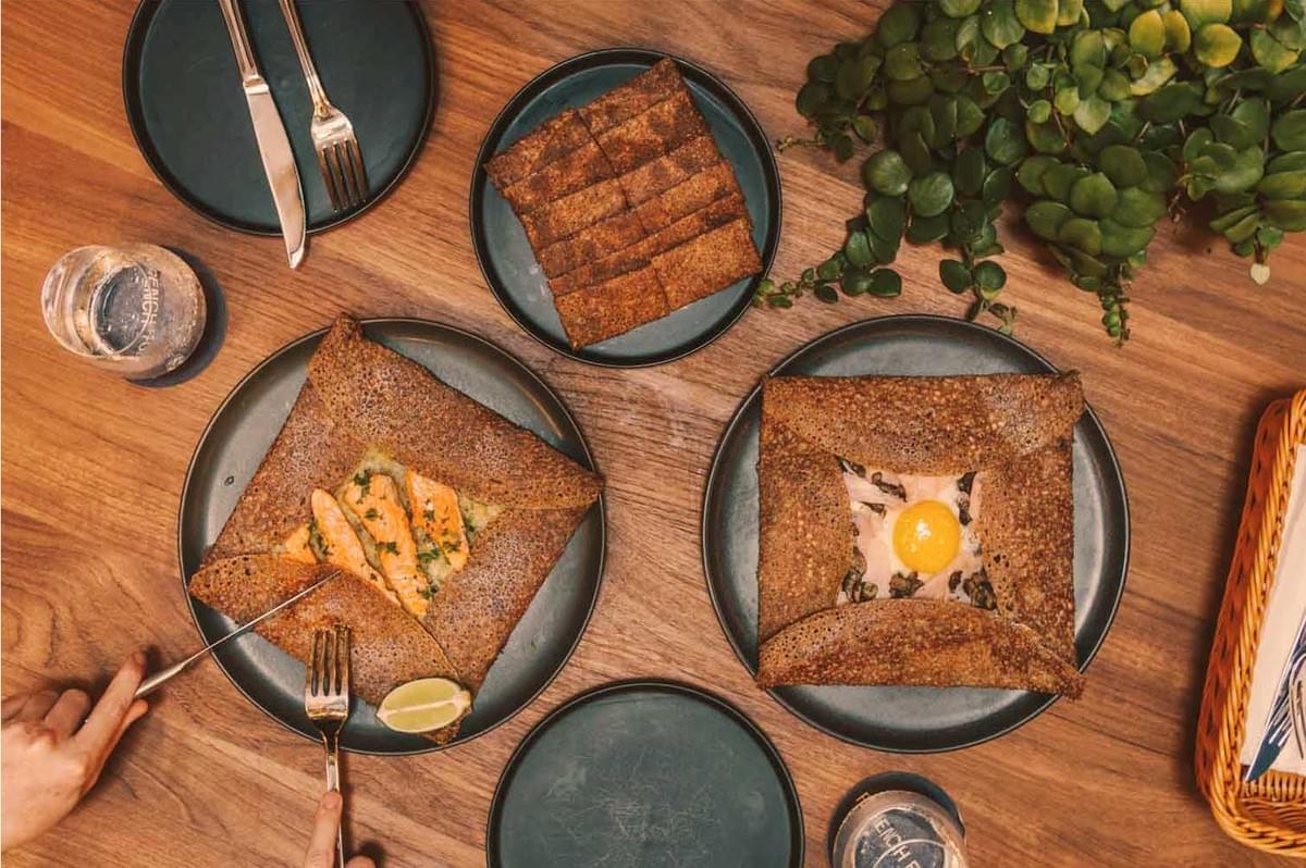 A wooden table with two plates of buckwheat galettes; one with salmon, the other with an egg. A plate of cut bread, a plant, and a hand holding cutlery are visible.