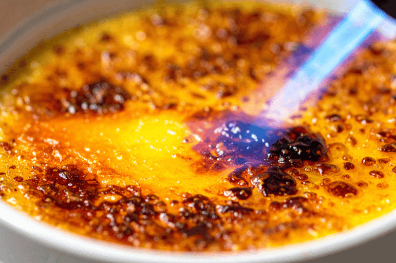 A kitchen torch flame being applied to a layer of sugar on a crème brûlée, showing the sugar melting and bubbling into a dark brown caramel.
