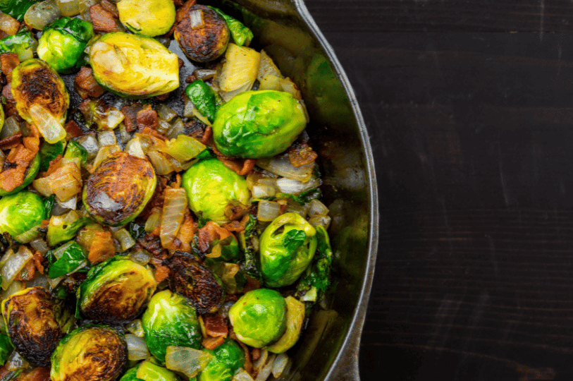 A top-down view of halved Brussels sprouts, diced onions, and crispy bacon bits searing in a seasoned cast-iron skillet.