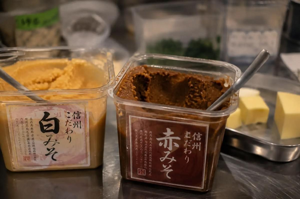 Two clear plastic storage tubs filled with Japanese fermented soybean paste sitting on a stainless steel work surface, with the tub on the left containing light-colored shiro miso labeled "信州こだわり 白みそ" and the tub on the right containing deep brown aka miso labeled "信州こだわり 赤みそ", accompanied by metal spoons resting inside each paste and cubes of butter on a small tray nearby.