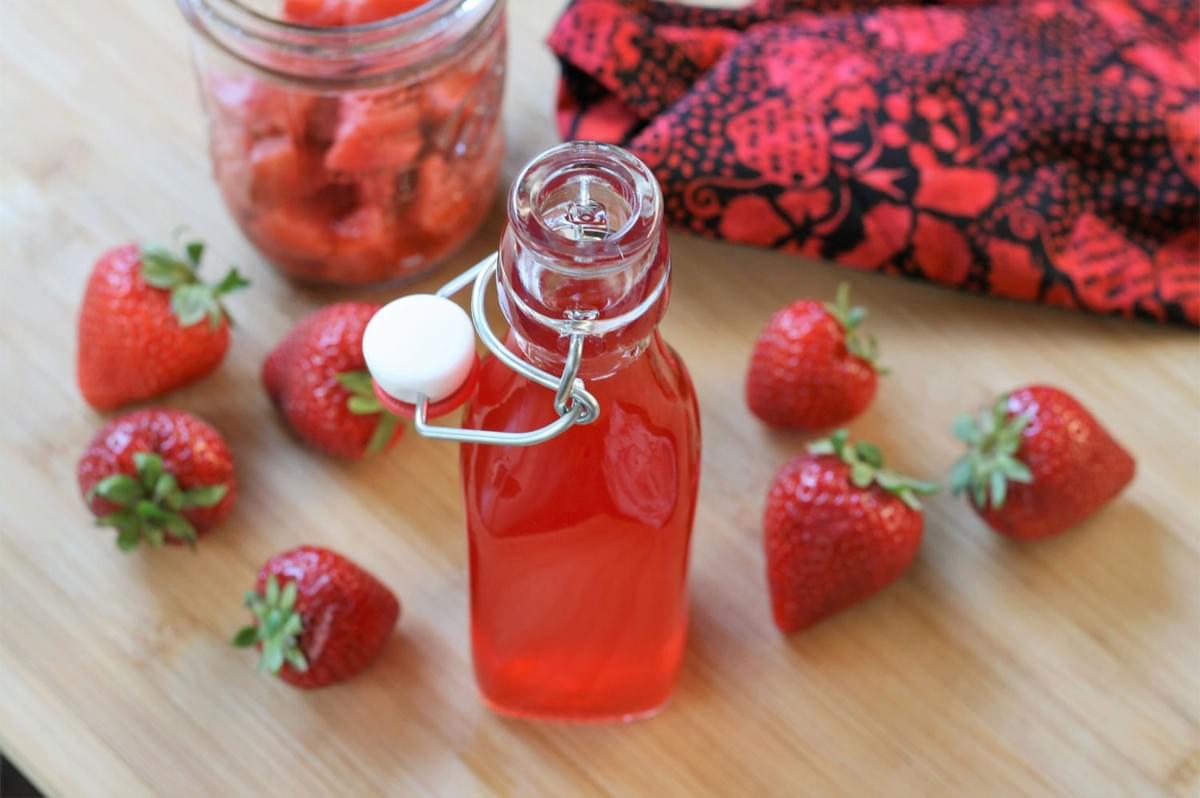 A small glass bottle with a swing top lid containing pink strawberry vinegar, surrounded by fresh strawberries, on a light wooden cutting board.