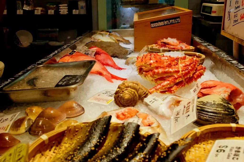 A seafood display at a market features a variety of fresh catch laid out on crushed ice, including a large red king crab, bright red fish, clams, and sea snails. Small price tags and bamboo baskets are arranged around the crushed ice bed in an indoor market setting.