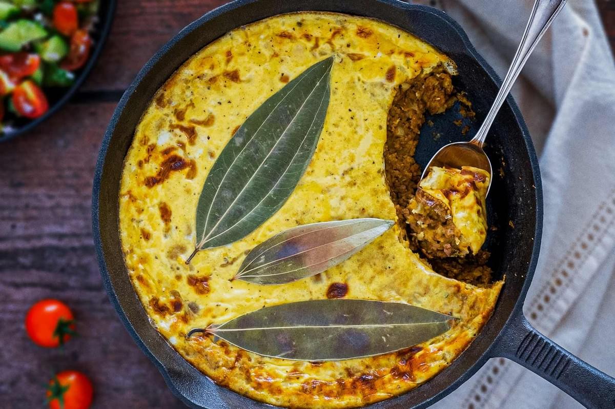 South African bobotie in cast iron skillet with golden custard topping, bay leaf garnish, and minced meat filling, served alongside fresh salad.