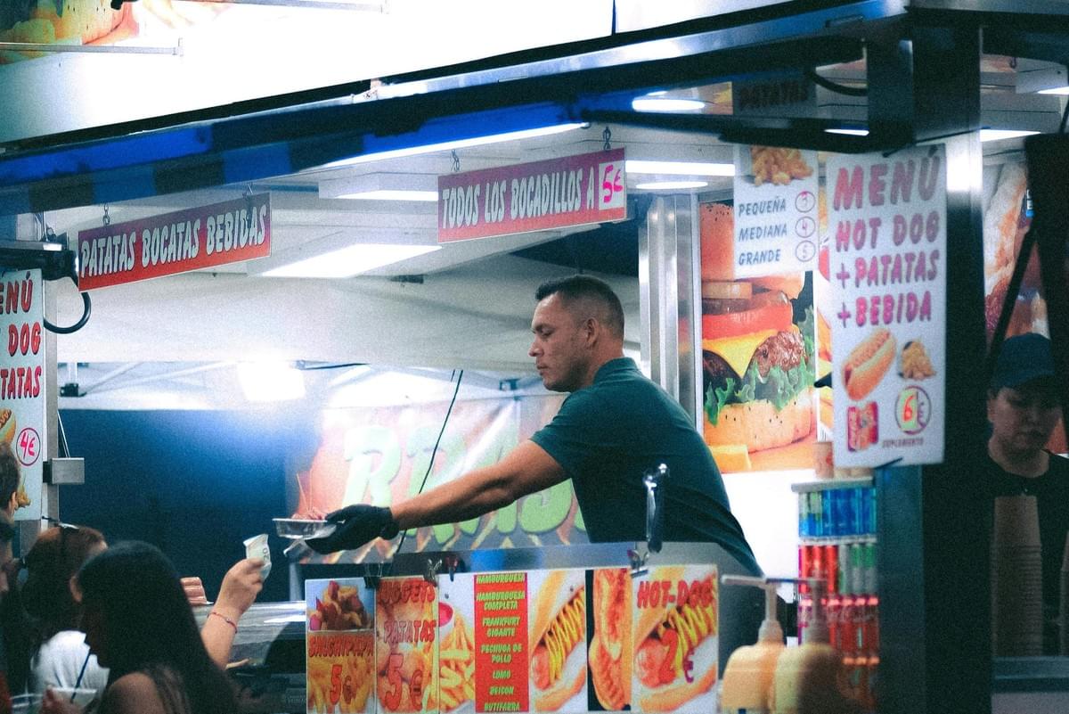 A vendor in a green shirt leans over the counter of a brightly lit food stall to hand a meal to a customer in exchange for cash. The busy stand is plastered with colorful signs in Spanish, advertising fast food options like hot dogs, sandwiches, and fries.