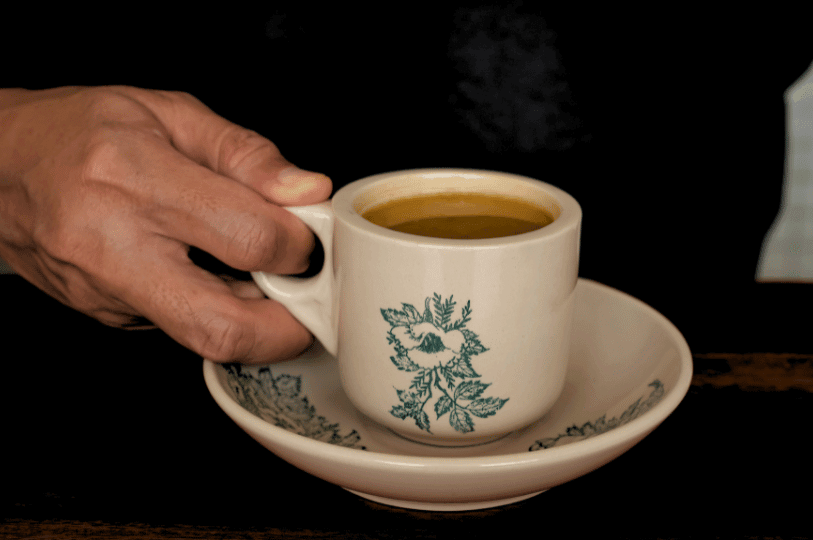 A hand reaching to grasp the handle of a traditional floral kopi cup containing a warm, milky coffee blend, set against a softly blurred dark background.