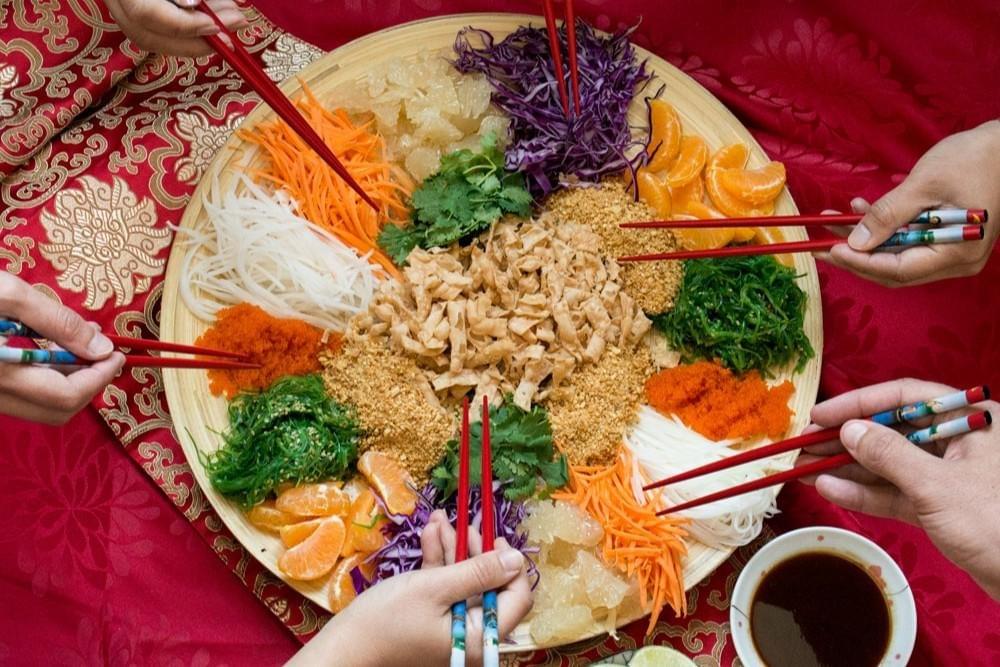  Gemini said Several people gather around a large circular platter to toss Yusheng, a colorful prosperity salad featuring shredded vegetables, citrus, and crispy crackers. The high-angle shot captures multiple hands using decorative red chopsticks to mix the ingredients over a vibrant red patterned tablecloth.