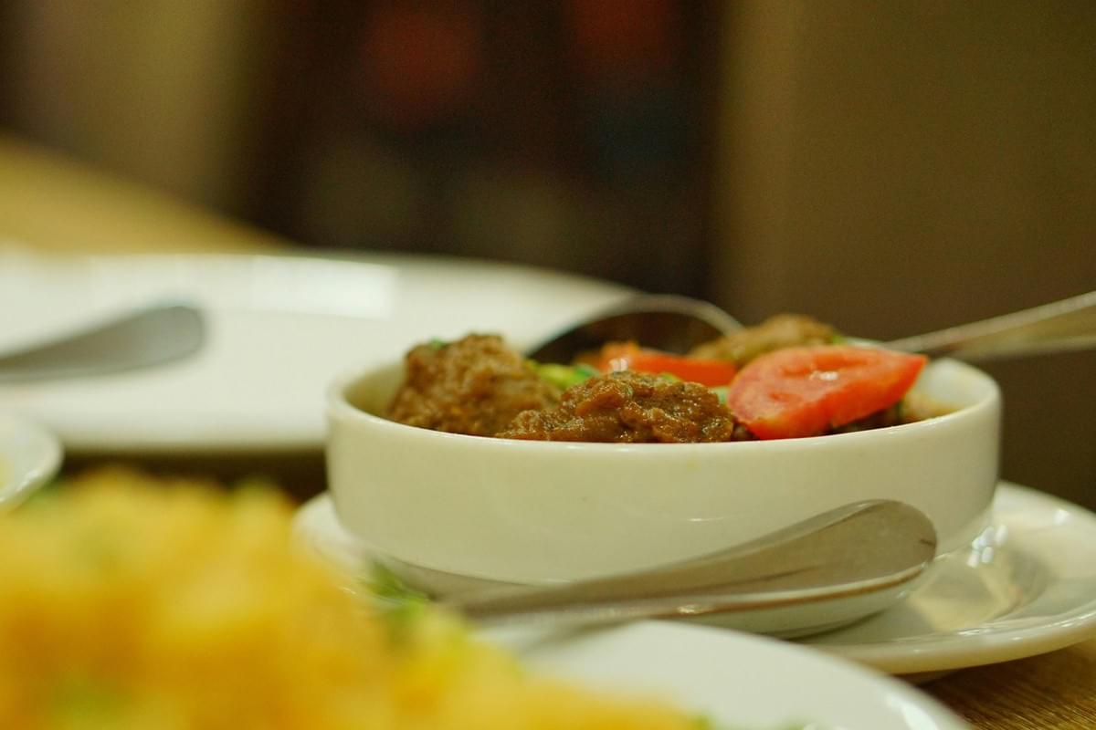 A shallow white bowl filled with a savory dish, featuring tender meatballs or chunks of meat in a thick sauce garnished with fresh tomato slices and green herbs. Metal serving spoons rest inside and beside the dish on a wooden dining table alongside other blurred plates.